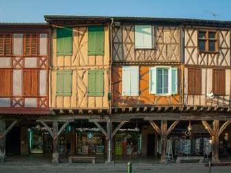 Bunte Fachwerkhäuser mit Holzbalken und Fensterläden in Mirepoix. Im Erdgeschoss befinden sich Geschäfte unter überdachten Arkaden.