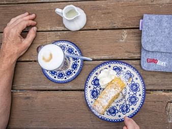 Apfelstrudel mit Puderzucker und Schlagsahne auf blau gemustertem Teller, Kaffeetasse und Eurobike-Tasche auf Holztisch während Radpause.