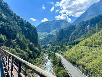 View from a wooden bridge into a forested mountain valley near Dogna. A turquoise river flows alongside a road through the valley.