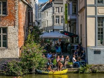 Menschen sitzen auf einer Café-Terrasse am Kanal in Gent. Gelbes Kajak im Wasser, historische Backsteingebäude säumen die enge Straße.
