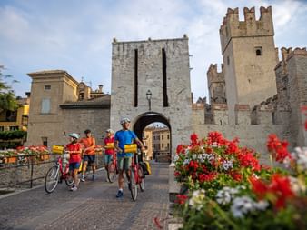 Four cyclists with bikes near medieval castle gate in Sirmione. Stone towers with crenellations frame an archway. Red and white flowers in foreground.