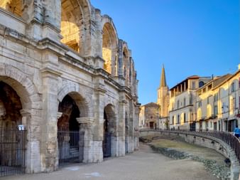 Ancient Roman amphitheater in Arles with stone arches and columns. Historic buildings and a church spire visible in the background under blue sky.