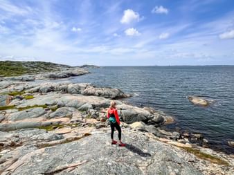 Cyclist in red jacket standing on rocky coastal landscape near Drottningviken, Amundön, overlooking calm blue sea under partly cloudy sky.