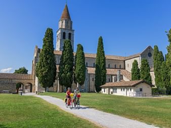 Three cyclists on a path leading to Aquileia Basilica with tall bell tower and cypress trees. Stone church complex under blue sky.