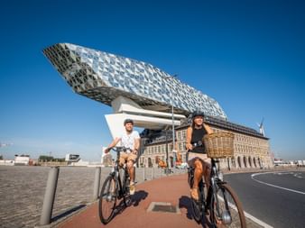 Zwei Radfahrer vor dem modernen Hafenhaus in Antwerpen mit seiner markanten diamantförmigen Glasstruktur auf einem historischen Gebäude unter blauem Himmel.