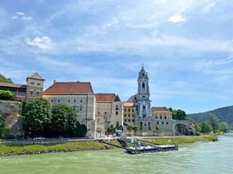 Stift Dürnstein mit blauem Barockturm am Donauufer in der Wachau. Historische Gebäude säumen das Ufer mit grünen Hügeln im Hintergrund.