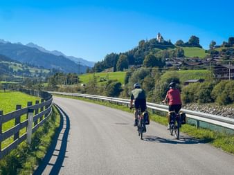 Two cyclists riding on a paved bike path with wooden fence, green meadows, and Alpine mountains. A church sits on a hilltop in the distance.