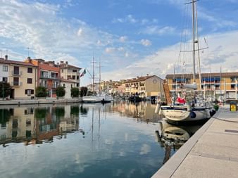 Ruhiger Hafenkanal in Grado mit vertäuten Segelbooten, bunten Gebäuden entlang der Uferpromenade und Spiegelungen im ruhigen Wasser unter blauem Himmel.