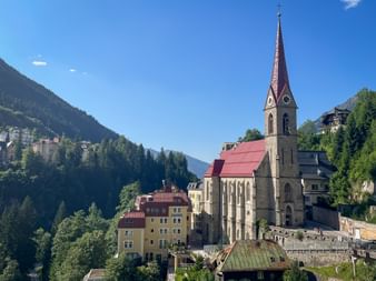 Parish church with tall spire and red roof in Bad Gastein, surrounded by colorful buildings and forested mountains under blue sky.