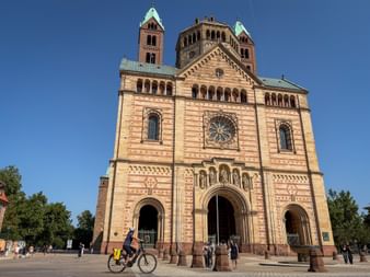 Radfahrerin vor dem Speyerer Dom, einer romanischen Sandsteinkirche mit Zwillingstürmen und grünen Kupferdächern unter blauem Himmel.