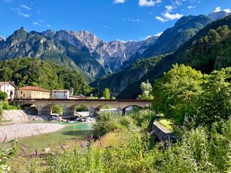 Steinbrücke über einen türkisfarbenen Fluss in einem Alpental mit Dorfhäusern und dramatischen Berggipfeln im Hintergrund unter blauem Himmel.