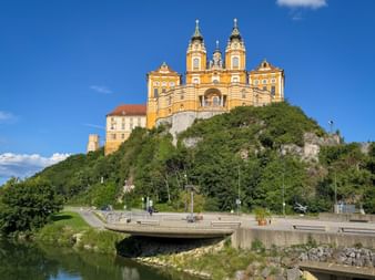 Gelbes barockes Stift Melk mit Zwillingstürmen auf grünem Hügel über der Donau. Eine Brücke überquert den Fluss im Vordergrund unter blauem Himmel.