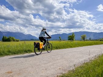 Cyclist with panniers riding on paved road through green fields near Murnauer Moos, with mountains and dramatic clouds in background.