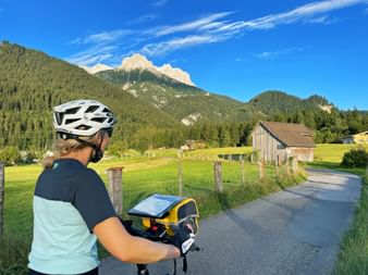 Radfahrerin mit Helm und Lenkertasche auf asphaltiertem Weg bei Saalfelden. Grüne Wiesen, Holzscheune und bewaldete Berge unter blauem Himmel.