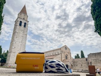 Gelbe Lenkertasche und weiß-blauer Fahrradhelm vor der Basilika Santa Maria Assunta in Aquileia mit Glockenturm und Zypressen.