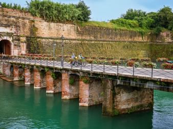 Two cyclists crossing a flower-decorated stone bridge over turquoise water, with ancient fortress walls and green vegetation in the background.