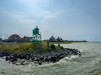 Green metal lighthouse on rocky breakwater at Stavoren harbor on IJsselmeer. Red-roofed buildings visible in background under blue sky.