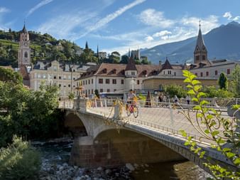 Cyclists crossing an ornate stone bridge over a river in Meran, with historic buildings, church towers, and mountains in the background.