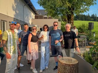 Group of eight people posing outdoors at a summer gathering, standing on a paved area with buildings and green countryside in background.