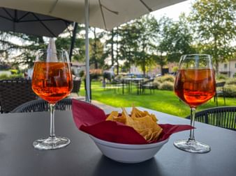 Zwei Gläser Aperol Spritz mit Eis und eine Schale Chips auf einem Tisch im Freien unter einem Sonnenschirm, mit Gartenblick in Friaul.