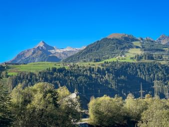 Alpine landscape near Kaprun with Kitzsteinhorn mountain, green slopes, forests, and a church tower under clear blue sky.