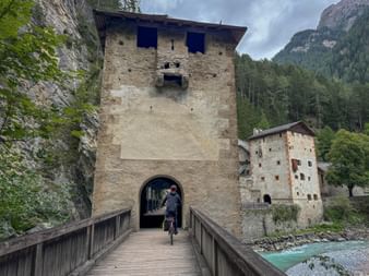 Cyclist crossing wooden bridge through medieval stone gate tower at Altfinstermuenz. Historic fortress walls and turquoise river visible below.