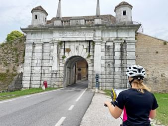 Radfahrerin mit weißem Helm betrachtet ein barockes Stadttor in Udine mit zwei runden Türmen und lateinischer Inschrift über dem Torbogen.
