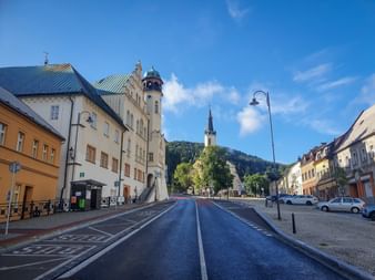 Wide street through Neudek market square with colorful buildings, clock tower, and church spire. Forested hills rise in the background under blue sky.