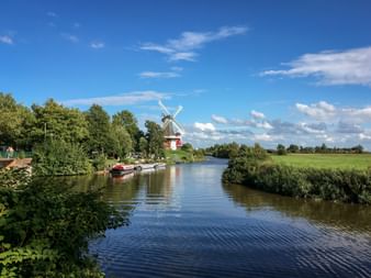 Historic twin windmills in Greetsiel along a peaceful canal with boats moored nearby, surrounded by green fields and trees under blue sky.