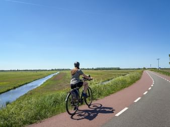 Radfahrerin auf rot gepflastertem Deichradweg entlang eines Kanals durch flache niederländische Polderlandschaft unter blauem Himmel.