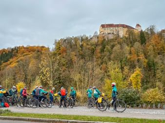 Group of cyclists in colorful jackets riding along Danube path in autumn. Castle on wooded hilltop with orange and yellow foliage.