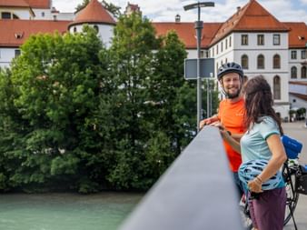 Zwei Radfahrer mit Helmen machen Pause auf einer Brücke in Füssen, Bayern. Historische weiße Gebäude mit roten Dächern und grünen Bäumen im Hintergrund.