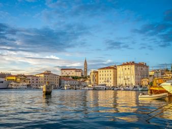 Hafen von Piran in Istrien mit Booten, historischen mediterranen Gebäuden und Kirchturm unter blauem Abendhimmel.
