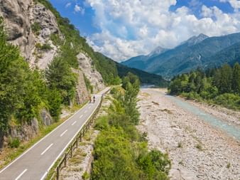 Two cyclists on paved path along turquoise river in Kanaltal valley, with rocky cliffs and forested mountains under blue sky with clouds.
