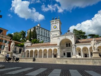 Weiße Renaissance-Loggia mit Uhrturm an der Piazza della Libertà in Udine. Zwei Radfahrer überqueren den Kopfsteinplatz unter blauem Himmel.