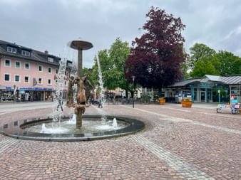 Richard Strauss Fountain with bronze sculptures in a cobblestone square in Garmisch, surrounded by colorful buildings and trees.