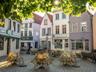 Kopfsteinpflasterplatz im Bremer Schnoorviertel mit bunten historischen Gebäuden, Café-Bestuhlung im Freien und grünem Laub.