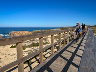 Two cyclists riding on a wooden boardwalk along the Atlantic coast in Alto Minho, Portugal. Sandy beach and rocky coastline visible beside the path.