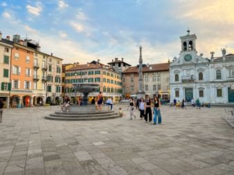 Piazza Giacomo Matteotti in Udine mit bunten historischen Gebäuden, weißer Barockkirche, zentralem Brunnen und flanierenden Menschen bei Sonnenuntergang.