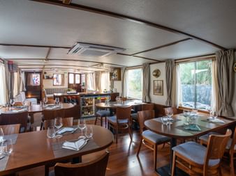 Innenraum des MS Caprice Salons mit hölzernen Esstischen, gedeckt mit Gläsern und Servietten, großen Fenstern mit Vorhängen und Barbereich im Hintergrund.