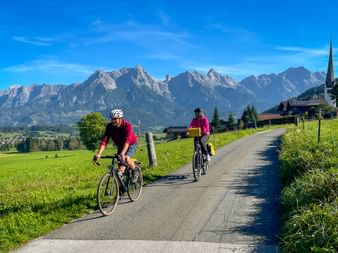 Two cyclists in pink jerseys riding on a paved road through green meadows with Alpine mountain range and church steeple in background.