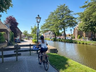 Radfahrerin fährt entlang einer Gracht in Schipluiden. Traditionelle niederländische Häuser und Bäume säumen den Wasserweg unter blauem Himmel.