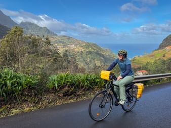 Female cyclist with yellow panniers on road near Boaventura, Madeira. Mountains, ocean, and scattered houses visible in background under blue sky.