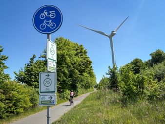 Radfahrerin auf asphaltiertem Weg mit Radwegschildern und Windrad. Grüne Vegetation säumt den Weg unter blauem Himmel bei Rotterdam.