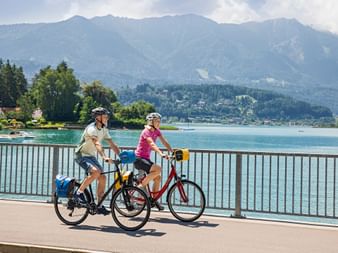Zwei Radfahrer auf einem Uferweg am Faaker See in Kärnten. Türkisfarbenes Wasser und bewaldete Berge im Hintergrund sichtbar.