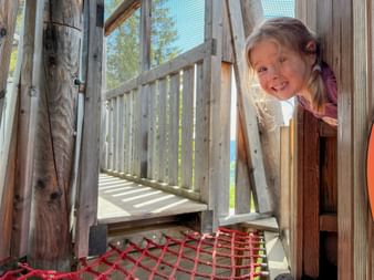 Smiling girl looking through window of wooden playground tower with climbing net and railing visible in sunny outdoor setting.