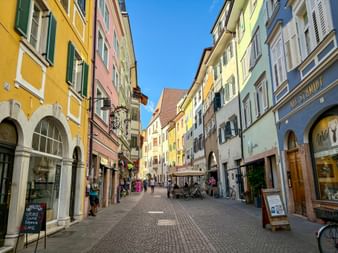 Pedestrian street in Bozen with colorful buildings in yellow, pink, and blue. Shops line both sides, people walk and sit at cafes under clear sky.