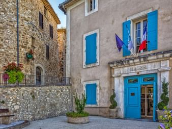 Steinernes Rathausgebäude mit blauen Fensterläden und französischen Flaggen auf einem provenzalischen Dorfplatz, umgeben von historischen Steingebäuden.