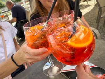 Two hands toasting with large glasses of Aperol Spritz with ice and orange slices at an outdoor café in Meran.