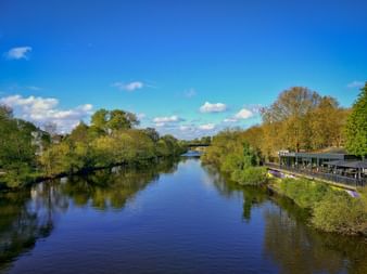 Friedliche Lahn fließt durch grüne Landschaft mit Bäumen an beiden Ufern, blauer Himmel mit weißen Wolken und Gebäuden in der Ferne.